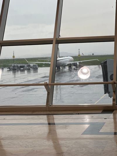 The image shows an airplane on a wet tarmac at an airport, viewed through large windows of a building. The tarmac is reflective, indicating recent rain. Several cargo containers and ground support equipment are visible near the airplane. In the background, there are distant hills or mountains and other airport infrastructure, including what appear to be navigation lights. The sky is overcast and gray. Inside the building, the floor appears to be tiled, and the light from a spotlight or runway light is visible reflecting on the window, creating a bright circular glow. The scene suggests a typical operational day at an airport, possibly during inclement weather. The specific airport is Bole International Airport in Addis Ababa, Ethiopia, given that Ethiopian Airlines is visible on the side of the aircraft.