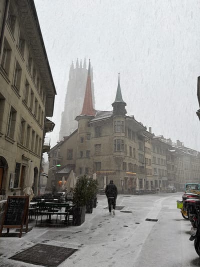 The scene depicts a snowy urban street in Fribourg - Freiburg, Switzerland, specifically the Rue de Lausanne facing towards the St. Nicholas Cathedral. Heavy snowfall is visible, accumulating on all surfaces and reducing overall visibility.

In the background, the gothic spire of the St. Nicholas Cathedral rises indistinctly through the snow-filled air, presenting a tall, intricate silhouette. In the mid-ground, a prominent multi-story building features a distinctive red conical roof and a smaller green conical turret at its corner, with bay windows. Below this building, storefronts are visible, including one marked "SCHWEIZERHALLE." To the left, a cream-colored stone building with multiple windows and balconies lines the street.

The street and sidewalk are covered in a fresh layer of snow, revealing numerous footprints, primarily leading away from the camera. Several dark manhole covers are embedded in the snow-covered ground.

One individual, dressed in a dark jacket, dark trousers, and white-soled shoes, walks away from the viewer in the center of the frame.

To the left, an outdoor cafe area is visible, furnished with dark metal tables and chairs, all dusted with snow. Several folded white outdoor umbrellas stand upright. A dark chalkboard menu sign, displaying "Menu du jour" and other text in white, stands on the pavement. Planters containing small green trees or shrubs are also present, covered in snow.

On the far right, the rear section of a dark motorcycle is partially visible, displaying a Swiss license plate "FR 40014." Adjacent to it, parts of a yellow bicycle-like object with a lime green bag and the rear of a light blue/green van with colorful artwork are discernible. A green-glowing traffic light or street lamp fixture extends over the street in the mid-ground. The overall environment is quiet and characterized by winter weather conditions.