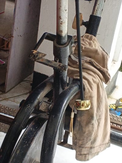The scene, located in Burauen, Philippines, depicts a close-up of a heavily worn, black metal frame, consistent with a utility bicycle or tricycle. The frame exhibits extensive dirt, scratches, and rust, particularly on its connection points and a prominent vertical support pipe, likely a seat post, which is painted white but shows significant paint loss and corrosion. A light brown, worn canvas-like fabric is draped over the upper-right section of the frame, displaying multiple folds. A golden-colored, reflective cylindrical object is attached to a thin, vertical black rod positioned parallel to the main frame. From this object, a light brown braided cord with a white toggle hangs downwards. Multiple curved black metal frame tubes are visible in the lower section, partially obscuring internal components, possibly a pedal crank or bottom bracket area. The background consists of a light-colored vertical wall and a floor with visible geometric patterns. No human subjects or direct interactions are present.
