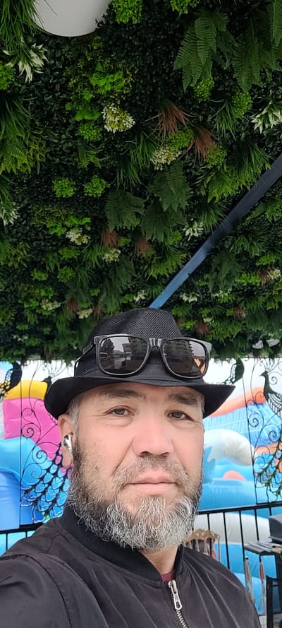 A man wearing a black fedora, black sunglasses, and a black jacket is pictured in front of a wall covered in artificial greenery. He has a salt-and-pepper beard and is wearing a white wireless earbud. The background behind him features a colorful inflatable structure with what appears to be peacocks or similar birds depicted on a fence. The man is looking directly at the camera. The scene is outdoors, likely in Milisi, Georgia, given the presence of an inflatable play structure often found at public gathering places or event venues.