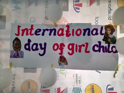 In Ajah, Nigeria, a white rectangular banner is prominently displayed against a backdrop of various corporate and organizational logos. The banner features the handwritten text "International day of girl child" in alternating red and purple lettering. Three cutout images of young girls are affixed to the banner: a smiling Black girl with her chin resting on her hands on the left, a White girl in a straw hat holding flowers on the right, and a smaller image of a White girl near the bottom center. Several inflated white balloons are positioned around the banner, attached to the display surface. The background logos, some inverted or sideways, include "Precious Schools", "WE MEDIY", and "ODIDO", among other geometric patterns, indicating an event or celebration setting.