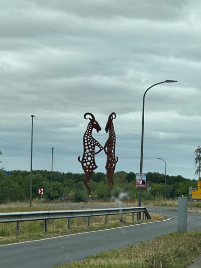 A metal sculpture of two spotted goats stands tall by a road in Bitburg, Germany. The goats are posed facing each other, their bodies adorned with numerous circular cutouts. A light pole is positioned behind the goats, and another one is visible to the left. In the background, a dense line of trees forms a backdrop against a cloudy sky. A guardrail runs along the side of the road. A traffic sign with a red and white chevron indicates a curve to the right. Further in the distance, a political poster featuring a man is attached to a pole. The scene is viewed from a vehicle, suggesting the photograph was taken while driving.