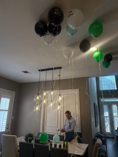 A residential interior in Southlake, Texas, United States of America, shows a person at a dining table with chairs, bottles, and balloons. The scene includes a multi-bulb light fixture, windows, and a decorative wall trim. Various balloons float overhead, some with graduation motifs. The person is dressed in a striped shirt and jeans and appears to be interacting with objects on the table. The overall environment suggests a celebratory gathering.
