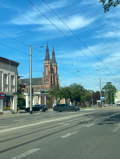 The image depicts a street scene in Pabianice, Poland, under a bright blue sky with scattered clouds. The perspective is from a moving vehicle, likely a car, indicated by the road, tram tracks, and overhead wires for the tram system. The orientation of the image is vertical, suggesting it was taken while turning or at an angle.
Dominating the background is a large, red-brick church with two prominent spires. The church appears to be an older, possibly gothic-style building. In the foreground and mid-ground, there are various buildings, some with visible storefronts. Trees line the street, providing shade and greenery.
Several cars are visible on the road, including a dark-colored sedan in the right lane and another partially visible car behind it. The activity is a typical urban street scene with vehicles and pedestrian infrastructure like tram lines and poles. The time of day appears to be daytime, given the clear sky and shadows. The weather seems pleasant.
Some text is visible on a storefront in the lower right portion of the image, including "PROPLAN" and "warta". The overall impression is a sunny day in a European city, with a mix of historical architecture and modern infrastructure.