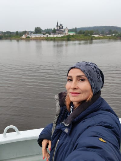 In the foreground, a woman with auburn hair and makeup is captured in a selfie from the chest up. She is wearing a dark blue winter jacket and a grey beanie with a pattern. The woman is standing on the deck of a vessel, as indicated by the white railing and a white loop fixture in the lower left corner of the image. In the background, across a body of water, the Goritsy Monastery is visible, a prominent landmark in Goritsy, Russia. The monastery features several white buildings with dark domes and a prominent bell tower. The environment appears overcast and the water is calm.