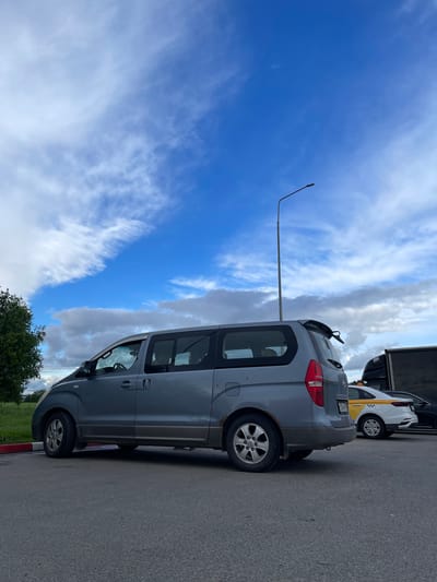The image shows a gray Hyundai Starex minivan parked on an asphalt surface next to a curb with red and white paint. To the right of the minivan is a white and yellow taxi, followed by the trailer of a truck. A single streetlight stands to the left of the minivan, reaching towards a blue sky with white clouds. A patch of green grass and trees is visible in the lower left corner. The location is identified as Бавыкино, Russia.