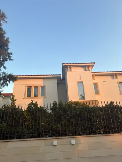A low-angle shot captures the upper floors of a modern, two-story villa in Pervolia, Cyprus, bathed in the warm, golden light of late afternoon or early morning. The villa's facade is a pale cream color, with red-tiled roofing visible. The building features prominent architectural details, including a stepped turret-like structure on the right side with decorative brackets under the eaves. Several rectangular windows are visible, some with reflective glass showing foliage. A decorative wrought-iron fence with ornate scrollwork runs horizontally across the foreground, partially obscuring dense, dark green bushes. Below the fence, a stone wall with horizontal block detailing forms the base of the scene. The sky above is a clear, pale blue, with a small crescent moon visible in the upper right quadrant. A portion of a dark green coniferous tree is visible on the far left of the image, its branches extending into the sky. The scene appears serene and residential.
