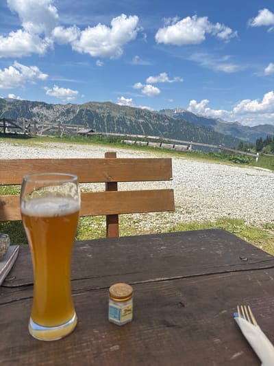 A glass of beer and a small jar sit on a wooden table. The table is in front of a wooden bench, which is in front of a gravel path. Beyond the path, a wooden fence runs along the side of a grassy slope. In the background, a mountain range with trees and rocky peaks rises against a blue sky with white clouds. The location is Ratschings - Racines, Italy.