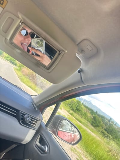 The image is taken from inside a car, focusing on the driver's side interior. The sun visor is flipped down, revealing a vanity mirror that reflects a person holding up a smartphone to take a selfie. The person is wearing sunglasses and sticking their tongue out. The dashboard and side mirror are visible, and through the side window, there's a glimpse of a road and green landscape. While Bulgaria is mentioned, the photo is not sufficient to confirm that the location is Balanovo, Bulgaria.