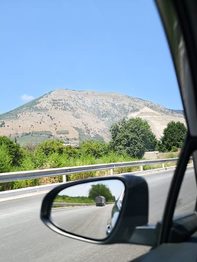 The image shows a view from inside a car on a sunny day. In the foreground, a side-view mirror reflects the road behind, showing a truck and trees. The car is on a road with a metal guardrail to the left. Beyond the guardrail, there is lush green vegetation and trees. In the background, a large, barren, light-colored mountain rises under a clear blue sky. The terrain of the mountain shows signs of terracing or past excavation, with some areas appearing lighter and disturbed. The overall scene suggests a rural or mountainous landscape, possibly in the vicinity of Nola, Italy, given the context of the prompt. No people are visible.