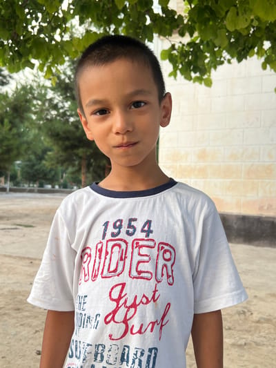 A young boy with dark hair and brown eyes looks towards the camera, his head tilted slightly. He is wearing a white t-shirt with a navy blue collar. The t-shirt has red and blue lettering that appears to say "RIDER 1954" and "Just Surf" in a stylized font. The boy is outdoors, with green foliage visible on the left side of the frame and a light-colored brick wall in the background. The ground is a light concrete or dirt surface. The lighting suggests it is daytime, possibly late afternoon given the shadows. The overall setting appears to be a casual outdoor environment, perhaps a yard or park in Balkh, Tajikistan. The boy's expression is neutral.