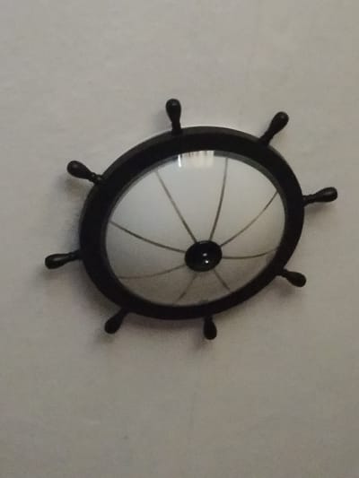 An artistic nautical-themed wall lamp, designed to resemble a ship's wheel, is affixed to a light-colored wall. The lamp has a dark, circular frame with eight spokes extending outwards, each tipped with a rounded handle. The center of the wheel features a glass dome, behind which a light source is visible. The overall impression is of a decorative light fixture with a clear maritime aesthetic. The image does not provide any information about the city of Osinniki, Russia.