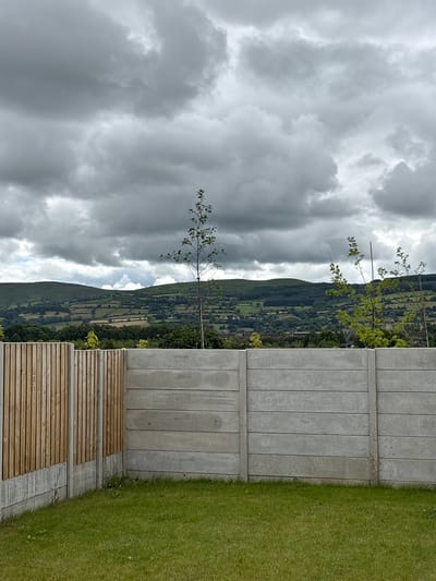 This is a vertically oriented, eye-level medium shot of a cloudy landscape in Clonmel, Ireland. The foreground is dominated by a modern concrete and wood fence, separating a manicured lawn from a view of rolling hills. The fence is constructed from horizontal wooden planks at the bottom and vertical concrete slabs above. The lawn to the right of the fence is vibrantly green and appears well-maintained.
Beyond the fence, a lush green hillside stretches into the distance, dotted with trees and fields, suggesting a rural or semi-rural setting. The sky is filled with dramatic, heavy grey clouds, indicating overcast weather. There are no people or specific activities depicted, creating a sense of stillness and natural beauty. The scene appears to be set during the daytime, with the diffused light suggesting it might be later in the afternoon or a particularly overcast day. The overall impression is one of a serene, natural environment juxtaposed with modern landscaping elements.