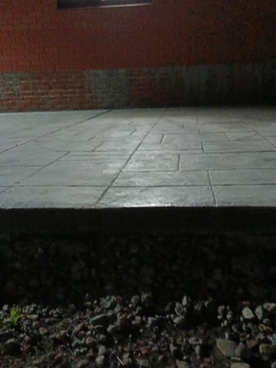 An outdoor scene at night shows a brick wall at the top of the frame, extending downwards into a concrete pathway. The pathway is made up of rectangular stones laid in a patterned arrangement and shows a reflection of light. Below the pathway, a dark, rough area is visible, appearing to be a bed of gravel with some sparse green foliage. The location is identified as Novotitarovskaya, Russia.