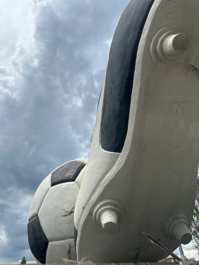 This is a low-angle, close-up shot of a large, cracked, white and black soccer ball sculpture, positioned next to a similarly oversized sculpture of a soccer cleat. The soccer ball is on the left, its black hexagonal panels showing wear and tear. The white cleat sculpture is on the right, its sole visible with three circular studs. The background consists of a cloudy, overcast sky. The scene appears to be outdoors, possibly in an urban or park setting, given the hint of a distant tree on the far right and a white structure at the bottom edge. The overall impression is one of weathered, monumental sporting equipment under a dramatic sky. The location is identified as Staryi Aidar, Ukraine.