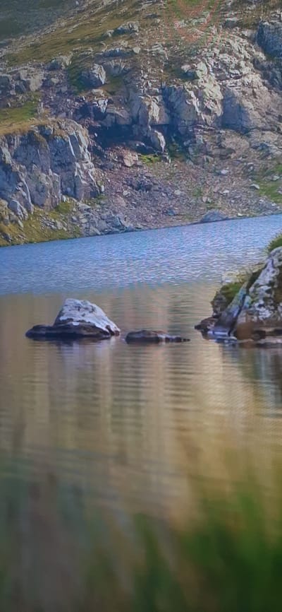 The image depicts a serene natural landscape featuring a calm lake and a rocky mountainside. The water in the foreground reflects the muted colors of the sky and the surrounding terrain, with subtle ripples disturbing its surface. Several dark, weathered rocks emerge from the water, breaking the uniformity of the reflection.

In the mid-ground, the lake stretches towards a steep, rocky slope. The mountainside is characterized by large, irregular rock formations, interspersed with patches of sparse vegetation. The terrain appears rugged and uneven, suggesting a remote, mountainous environment. The upper portion of the image shows more of the rocky slope, with a gradient of gray and brown hues dominating the rocky surfaces, and streaks of green indicating plant life clinging to the crevices.

The overall impression is one of a quiet, natural setting, likely a mountain lake in a region with significant geological activity. There are no people or man-made structures visible in the image. The image does not provide any information that would associate it with the city of Аксеново, Russia.