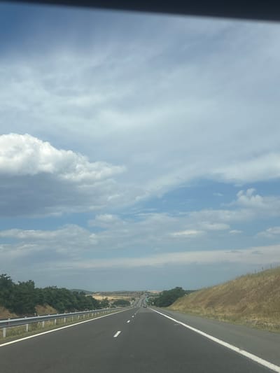 An eye-level, medium shot captures a straight, multi-lane highway receding into the distance under a cloudy sky. The road's asphalt surface is dark and marked with white lane dividers. On the left, a metal guardrail runs alongside the road, bordered by green trees. The right side of the highway features a dry, grassy embankment. In the distance, more trees and rolling hills are visible, with a few cars on the road. The sky is filled with a mix of white and gray clouds, with patches of blue sky peeking through.