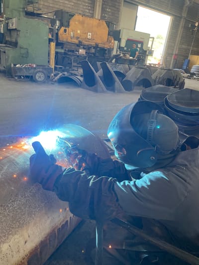 In an industrial setting, a person is welding a large metal cylinder. The individual wears protective gear, including a welding helmet and gloves. Sparks and bright light emanate from the welding point. In the background, there's machinery, metal components, and another person visible in the distance through a window. The location is Zaljevo, Montenegro.