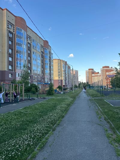 In Alexandrovka, Russia, a wide concrete pathway stretches into the distance, flanked by a lush green lawn dotted with white clover flowers. On the left, a multi-story residential building with a curved facade and large blue-tinted windows dominates the scene. Adjacent to the building is a playground with various structures, including monkey bars and swings. Several children are visible on the playground. Further down the pathway, a young woman in a white shirt and gray pants walks away from the camera. On the right side of the path, a fenced-in sports court is visible with a few people present. The sky above is a clear blue with a few wispy clouds, and power lines crisscross the upper portion of the image. The overall setting appears to be a modern residential area.