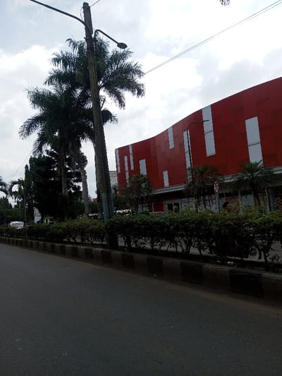 The image depicts a street scene in Khajuripada, India, under an overcast sky with scattered clouds. In the foreground, an asphalt road runs horizontally, bordered by a curb featuring alternating black and white painted sections. Beyond the curb lies a planted median strip dense with green shrubbery. A tall, dark street light pole stands prominently in the left mid-ground, extending upward with an arm and light fixture directed to the right. Several large palm trees and other dense trees are visible behind and to the left of this pole. On the right side of the frame, a large building with a red facade is visible. This facade is accented with irregularly spaced white vertical rectangular panels and features a wavy upper profile. The building's ground level includes visible windows and entrances. Partially obscured by the shrubbery near the red building, a male individual is visible, appearing to operate a two-wheeled vehicle, possibly a motorcycle or bicycle, with a portion of the vehicle's wheel visible beneath the shrubs. Overhead utility lines are present across the sky.