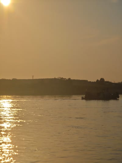 A wide shot captures a body of water bathed in the warm, golden light of a setting sun. The sun, partially visible in the upper left corner, casts shimmering reflections across the water's surface, creating a pattern of light and shadow. In the distance, a silhouetted shoreline rises, dotted with trees and a single tower-like structure. A barge, also silhouetted, navigates the water from right to left. The sky is a pale orange, with soft clouds present. The scene appears to be a natural landscape. It is not possible to determine the specific location within Lesosibirsk, Russia, from the visual information provided.