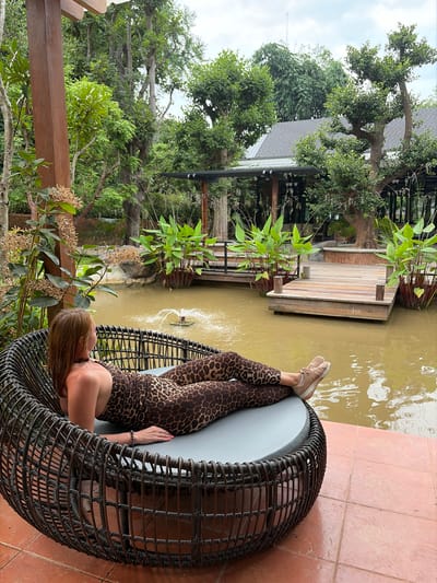 A woman with auburn hair, wearing a leopard-print jumpsuit and beige sneakers, is relaxing in a dark, woven chair. She is facing away from the camera, looking out at a murky body of water with a small fountain. Behind the water is a wooden deck and a building with a gray roof, surrounded by lush green trees and foliage. The scene appears to be an outdoor seating area of a resort or restaurant in Mae Rim, Thailand, suggesting a tropical or subtropical environment. The lighting is overcast, indicating a cloudy day.