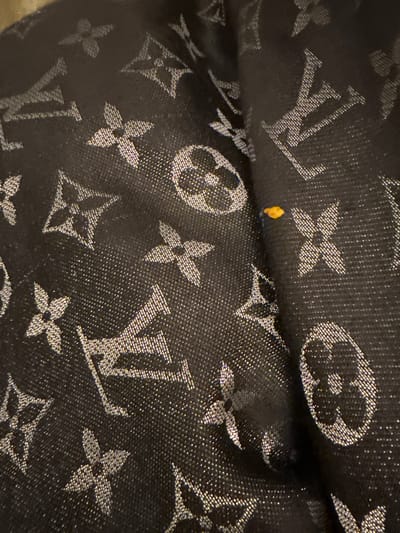 A close-up shot shows a repeating Louis Vuitton monogram pattern in silver-grey against a dark background, possibly fabric. The iconic "LV" initials and floral quatrefoils are densely scattered. A small, yellow speck is visible amidst the pattern, possibly a stain or debris. The material appears textured, with fine grain or weave details. The focus is sharp, highlighting the intricacies of the print.