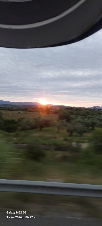 A wide shot captures a blurry landscape as seen from a moving vehicle, likely a car, traveling on a road. The foreground features a grey metal guardrail, indicating the edge of a roadway. Beyond the guardrail, the scene depicts a rural environment with blurred green foliage, including olive trees, and a field. In the background, rolling hills or mountains are visible under a cloudy sky. The sun is setting or rising, casting an orange glow on the horizon and illuminating the sky with soft, diffused light. The image is dated "9 юни 2026 г. 06:27 ч." and shows "Galaxy M52 5G" as the device used to capture it. The location is identified as Pont de Molins, Spain. There are no people or discernible subjects in the frame, only the natural landscape and the artificial road barrier. The motion blur suggests the photograph was taken while in transit.