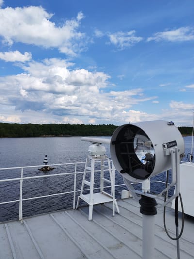 The image shows a view from the deck of a ship on a river, possibly in the vicinity of Novodevich'e, Russia, although this cannot be definitively confirmed from the image itself. The foreground features a large, white spotlight fixture, mounted on a pedestal, and a white radar or antenna structure. A white railing runs along the edge of the deck.

Beyond the railing, the river stretches to the horizon, bordered by a treeline. In the river, there is a small rocky islet marked with a black and white navigational beacon. The sky is bright blue with scattered cumulus clouds.