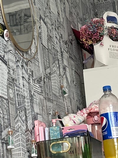 The image shows a cluttered tabletop with various items. On the left is a bouquet of dried flowers in a paper wrapping. Next to it is a plastic bottle of Pepsi. A variety of objects, including what appear to be makeup items (a pink compact and a green makeup case), are stacked. The background is a patterned surface, possibly a tablecloth or wallpaper, depicting a city aerial view. The location is not possible to determine from this image.