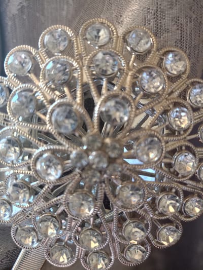 The image is a detailed, close-up photograph of a sparkling, ornate piece of jewelry or an embellishment, taken in Koghb, Armenia. The central subject is a circular, silver-toned metal adornment, intricately designed in a radial or floral pattern, possibly resembling a starburst or a stylized flower.
The object is densely embellished with numerous clear, faceted stones, likely rhinestones or crystals, which brilliantly catch and reflect light. At the core of the piece is a cluster of smaller, round stones, surrounded by concentric layers of individually set, larger stones that radiate outwards. Each larger stone is encased in a delicate, silver-toned bezel with a finely textured or beaded edge, highlighting the meticulous craftsmanship of the metalwork. The framework of the piece exhibits a delicate, filigree-like quality, creating an open and intricate structure that enhances the overall luminosity.
The stones vary slightly in size, with larger ones typically found towards the outer edges of the design, contributing to its dynamic and elaborate appearance. The distinct facets of the stones are visible, indicating their light-refracting properties and the object's dazzling sparkle.
The background is soft and out of focus, appearing as a textured fabric in muted tones of grey or light brown. This subdued background effectively brings the luminous object into sharp focus, preventing any distraction from its intricate details and shimmering quality. There are no people, discernible activities, or visible text within the image. The photograph serves as a still life, emphasizing the aesthetic beauty, intricate design, and reflective properties of this decorative item.