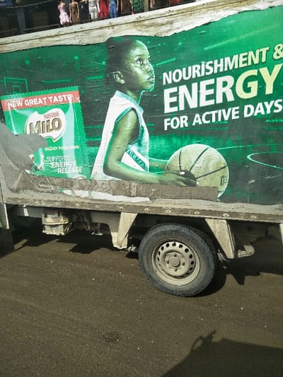 This image displays the side of a dirty, off-white commercial truck, positioned on a dark asphalt surface in Ajah, Nigeria. The truck's cargo bed is adorned with a large, weathered advertisement for Nestlé Milo, predominantly green and white. The advertisement features a young, dark-skinned female, depicted from the waist up, wearing a light-colored sleeveless jersey and holding a basketball. She is looking upwards and to the right. Visible text on the advertisement includes "NOURISHMENT & ENERGY FOR ACTIVE DAYS," "NEW GREAT TASTE," and "SUPPORTS ENERGY RELEASE." The advertisement is wrinkled and torn in several places, particularly on the left side where a depiction of a Milo packet is partially obscured. Below the cargo bed, the truck's chassis, undercarriage components, and a single rear wheel with a Bridgestone tire on a light-colored steel rim are visible. The asphalt ground appears textured and possibly damp or dusty. In the blurred background above the truck, various colorful garments are hanging, indicative of a street market or retail display. No live individuals are present within the immediate foreground or midground of the scene.