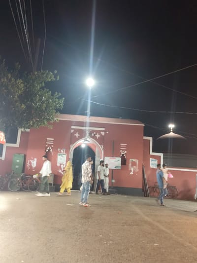 The image depicts a nighttime scene outside a red-colored building, possibly a religious site, in Bagerhat, Bangladesh. The building has a large arched entrance and decorative white elements on its facade. A dome with a finial is visible on the right side of the building. Several individuals are present, some walking and others standing. One person is carrying a child. Bicycles are parked near the building on the left. The ground is a paved surface. Overhead, power lines are visible against the dark night sky, with a bright light illuminating the scene from the right. The atmosphere appears to be one of a public gathering or a place of worship.