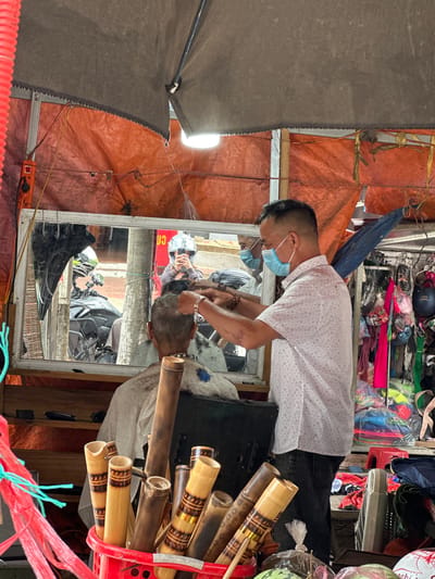 The image depicts an outdoor barbershop in Bắc Yên, Vietnam. A barber, wearing a light-colored, patterned short-sleeved shirt and dark jeans, is giving a haircut to a man sitting in a chair, who is covered with a white cape. The barber is wearing a light blue face mask. A mirror behind the barber reflects the street scene, showing a motorcycle and a person wearing a helmet. The barbershop appears to be set up under a makeshift roof covered by an orange tarp, with a gray tarp overhead. In the foreground, a red basket holds several bamboo tubes, some decorated with dark bands. Various items, including bags of merchandise, are visible in the background. The overall setting suggests a busy, informal commercial area.