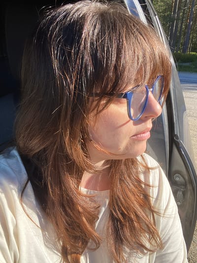 A woman with long, brown hair and blue-rimmed glasses sits in the passenger seat of a car. She is looking out the open car door at a sunlit forest landscape. The car is parked on the side of a paved road. The woman is wearing a white, long-sleeved shirt. The lighting suggests it is either early morning or late afternoon.