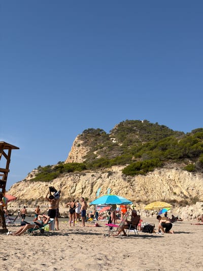 This is a wide, eye-level shot of a crowded beach on a sunny day. The sky is a clear, bright blue. In the foreground, golden sand is visible with several people lounging on chairs, lying on towels, or walking. One person is sitting in a lounge chair, reading a book, while another is kneeling on the sand. In the midground, a group of people are standing and interacting, with some changing clothes or talking. Beach umbrellas, one bright turquoise and one yellow, provide shade for beachgoers. In the background, a steep, rocky cliff face covered in green vegetation rises up, with some buildings visible near the top. A wooden lifeguard tower stands to the left of the frame. The beach appears to be in Urbanització Cales I Talaies, Spain, given the style of the buildings and vegetation.