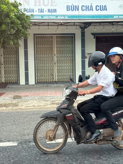 The image depicts a motorcycle with two individuals in motion on a street in Triệu Phong Commune, Vietnam. The rider, a young male wearing a white collared shirt, black pants, and a black helmet, is operating the motorcycle. He has a yellow wristband on his left wrist. Behind him, a female passenger wears a black jacket and a white and blue helmet. The motorcycle itself is black with red accents and has the "YAMAHA" logo visible. In the background, a building with metal roll-up doors is visible, and signage in Vietnamese indicates business names, including "BÚN CHẢ CUA" and what appears to be location-related text like "GÂN - TÁI - NAM" and "THƯỢNG - TRIỆU PHONG - QUẢNG TRỊ". The street is paved, with a white line marking a lane. The overall scene suggests a typical street view in a Vietnamese town or city.
