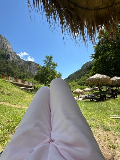 The image captures a serene outdoor scene in Zgorigrad, Bulgaria, likely from a seated perspective under a thatched umbrella. In the foreground, the lower half of a person's legs, clad in light pink pants, dominate the view. The background unfolds into a lush green landscape with a river meandering through it. Several straw umbrellas and lounge chairs are set up on the grass, suggesting a recreational area. Beyond the grassy area, a gently sloping hillside covered in trees rises, leading up to imposing, rugged mountains that form the backdrop. A small bridge with red and white railings is visible on the left side of the frame, spanning across a grassy incline. The sky is a clear, bright blue with a few wispy white clouds. The overall impression is one of tranquility and relaxation in a natural setting.