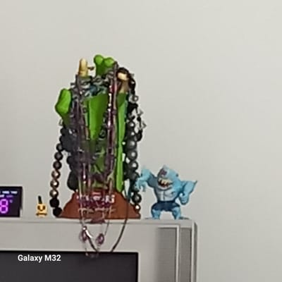 The image shows a close-up view of a shelf or surface, with several objects on it. To the left, a digital display shows the number "8" in purple digits. Next to the display is a small, gold-colored object that resembles a stylized figure or charm. The central focus is a green, sculpture-like object draped with multiple strands of dark, beaded necklaces. To the right of the green sculpture, a light blue toy figure of a muscular, shark-like creature is standing. The background is a plain, light-colored wall. In the foreground, a portion of a television screen is visible, with the text "Galaxy M32" superimposed in white letters at the bottom. The scene appears to be indoors, likely in a domestic setting. There is no discernible information in the image to indicate the city of Olsztyn, Poland, or any specific location.