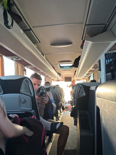 The image shows the interior of a bus, possibly in transit. Several passengers are visible in various states of relaxation, some sitting and looking at their phones. The bus interior features overhead compartments and seating. The location is in Bošnjaci, Croatia.