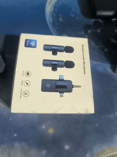 The image shows a cardboard box with a depiction of a wireless lavalier microphone system. The system consists of two microphones and a transmitter. The box is sitting on a surface, with the box's background being out of focus. The text on the box says "Wireless Lavalier Microphone". The location at the given coordinates is in Sapele, Nigeria. The image provides no information about people or any actions.