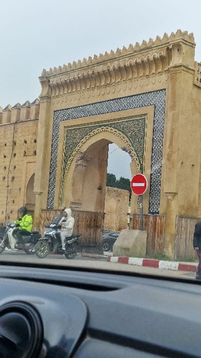 The scene depicts an urban street in Mechouar Fes Jdid, Morocco, viewed from the interior of a vehicle's dashboard. A large, ornate archway is central, constructed from weathered tan stone or stucco, featuring intricate dark and white geometric tilework (zellige) framing the opening and along the arch's curve. The arch's interior reveals exposed brickwork. To the left of the arch, a crenellated wall of similar construction exhibits evenly spaced small apertures. Beyond the arch, through the passage, distant green trees and a lighter, overcast sky are visible.
Two individuals on scooters are positioned on the street in front of the archway. The rider on the left wears a bright yellow-green jacket and a dark helmet, seated on a light-colored scooter. The rider in the center wears a white hooded garment and a white helmet, operating a dark-colored scooter. Their actions indicate they are either stopped or moving slowly. On the far right, a pedestrian's lower right leg and foot, clad in dark pants and patterned shoes, are partially visible on the sidewalk. A red circular "No Entry" sign with a white horizontal bar is mounted on a pole on the right sidewalk, adjacent to a light-colored concrete barrier. The curb along the street is marked with alternating red and white stripes. A dark sedan is partially visible behind the central scooter.