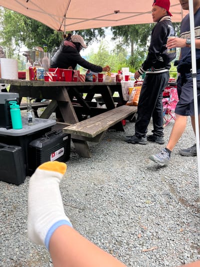 This image depicts an outdoor gathering, likely a picnic or barbecue, taking place under a portable canopy tent. The ground is covered in gravel, with a wooden picnic table as the central focus. On the table, there are various items including red cups, bottles of beverages, a large pot, and bags of snacks. Several individuals are visible: one person in a black jacket and white cap is attending to items on the table, another person is wearing a red baseball cap and a black jacket with white stripes, and another individual is wearing a dark blue t-shirt with white stripes. A person's leg with a white and blue sock is prominently featured in the foreground, suggesting a first-person perspective or a deliberate focus on the ground level. The environment suggests a natural setting with trees in the background. While the image does not explicitly state the location in Chilliwack, Canada, the presence of a portable canopy and picnic table suggests a park or recreational area commonly found in such a city.