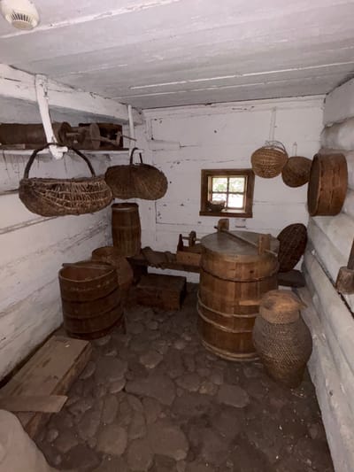 This is a photograph of the interior of a traditional, rustic building, likely a historical exhibit or a preserved dwelling, located in Sielachowskie, Poland. The walls and ceiling are constructed from rough-hewn wooden logs and planks, painted white with visible signs of age and wear. The floor is covered with irregularly shaped stones.

Dominating the center of the room is a large, wooden vat with a metal lid and two wooden paddles resting on top. To the left of the vat are two cylindrical wooden barrels. Several woven baskets of various sizes and shapes are displayed throughout the room. Two are hanging from the ceiling or upper wall, one is leaning against the wall to the right of the large vat, and another is situated in front of the vat and to its right, appearing to be a large, lidded basket. Smaller woven items, possibly bowls or hats, are also visible.

On a wooden shelf above the barrels, a piece of agricultural equipment, possibly a manual grinding tool or a flax-spinning device, is displayed. A small, square window with multiple panes is present on the back wall, allowing a glimpse of greenery outside. The lighting is dim, creating shadows and highlighting the texture of the wooden elements and the stone floor. The overall impression is one of a storage area for historical agricultural or household items. There are no people visible in the image.