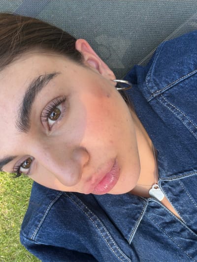 A close-up, upward-angled view of a person's face. The person has light skin, dark eyebrows, and brown eyes. They are wearing a dark blue denim jacket and silver hoop earrings. A silver necklace with a heart-shaped pendant is visible. The background appears to be a mix of a gray textured surface and green grass. The location in Santa Ana Yancuitlalpan is not discernible from the image.