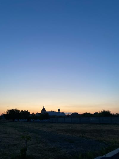 A vertical, wide-angle shot captures a twilight scene over Voznesensk, Ukraine. The sky transitions from a deep, clear blue at the top to lighter shades of blue and pale yellow near the horizon, indicating the end of sunset. Silhouetted against the horizon are several structures, including what appears to be a church with a prominent dome and cross, flanked by other buildings with pitched roofs. To the left, a large, dark tree stands out. In the foreground, a dry, grassy field is visible, with a small, thin sapling growing in the center. A concrete fence runs horizontally across the middle ground, adorned with a white, decorative pattern and some graffiti, including the words "ILoveYou." The overall impression is of a quiet, rural or suburban landscape at dusk.