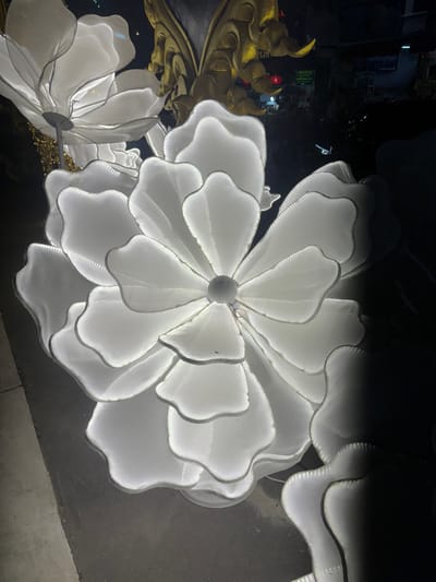 The scene depicts an outdoor nighttime view in Chiang Rai City Municipality, Thailand. The primary subjects are multiple large, white, illuminated floral sculptures. The central sculpture showcases layered, translucent petals with internal white lighting accentuating their undulating edges and structural lines. A similar, partially visible sculpture is situated to the upper left. Behind these, golden, ornate architectural elements, consistent with traditional Thai design, are partially visible, accompanied by hanging golden decorative strands. The far background on the right reveals faint urban lights, including small red lanterns and a green sign displaying white Thai text, confirming an urban environment. The sculptures rest on a dark paved surface. No individuals or active events are present.