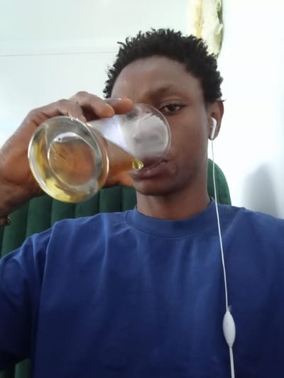 The image shows a young Black man wearing a blue t-shirt and earphones. The man is seated in a chair with a green back, and he is holding a glass of liquid with his right hand, bringing it up to his mouth to drink. The background is white, and there is a partial view of a white ceiling fan in the upper right of the image. The scene is in a residential setting in Ujevwu, Nigeria. The location is approximated based on the provided coordinates (5.471, 5.818), which are located in the city of Ujevwu, Nigeria.