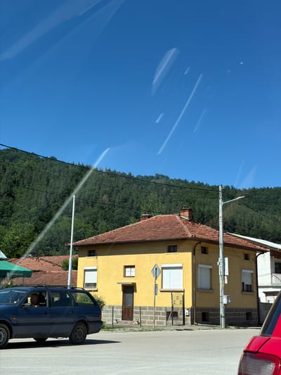In Beli Osam, Bulgaria, under a clear blue sky, a bright yellow house with a terracotta tiled roof stands prominently. The house has several windows, some with white rolling shutters, and a wooden door. A street lamp is affixed to a utility pole near the house. In the foreground, a dark blue station wagon is parked, with a driver visible inside. A partial view of a red vehicle is also present on the right side of the image. Behind the house, a dense green forest covers a hillside, with a thin white stream of water arcing across the sky from the left, originating from a pipe extending from beyond the frame and falling towards the hillside. The scene appears to be a residential area with a natural backdrop.
