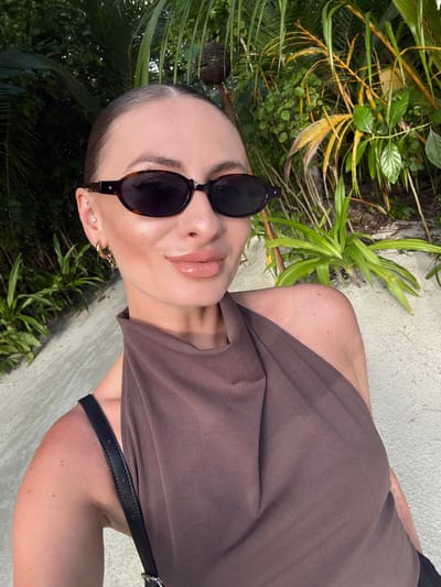 A woman with her hair pulled back, wearing sunglasses and a brown halter top, poses for a selfie outdoors. She has on light-colored lipstick and visible earrings, including a hoop earring and studs in her left ear. The background features lush green foliage, including palm trees and other tropical plants, suggesting a tropical environment. The ground appears to be sandy or a light-colored pathway. The overall setting is suggestive of a resort or natural landscape in the Maldives.